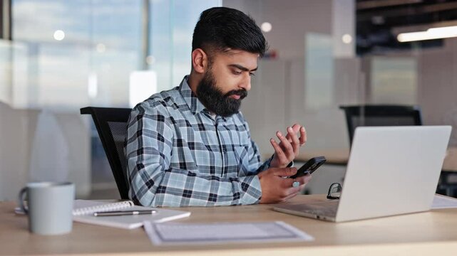Bearded businessman working in modern bright office at desk, using phone. Frustrated and young man in glasses user feel angry dissatisfied with incorrect slow software work.
