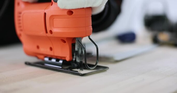 Carpenter hands in gloves skillfully operate orange jigsaw cutting wooden parquet panel. Worker makes hole in plywood board win carpentry shop