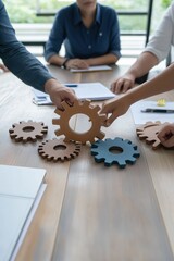 A group of people are holding gears together on a table. The gears are made of wood and are arranged in a circle. The people are working together to create a machine or a device