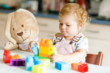 Adorable toddler girl with favorite plush bunny playing with educational toys in nursery. Happy healthy child having fun with colorful different plastic blocks at home. Cute baby learning creating.