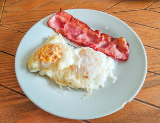 Crispy fried eggs and a strip of perfectly cooked bacon on a simple white plate.  A classic breakfast food image, perfect for menus, blogs, or culinary websites.  Highquality, appetizing photography.