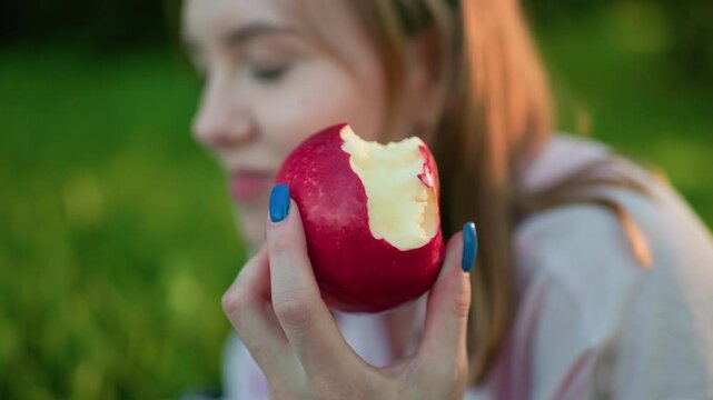 Close up of young woman holding a bitten red apple in her hand with blue-painted nails, smiling softly in a grassy field, enjoying nature and relaxation, as sunlight filters through surrounding