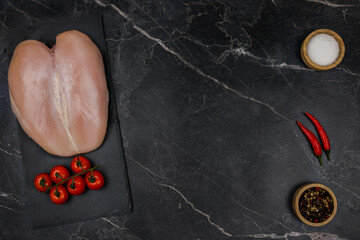 Obraz premium Chicken breast fillet with tomato sprig lies on a black board on the other side with spices and salt in wooden bowls and red hot pepper on a dark background with space for text in the center. The