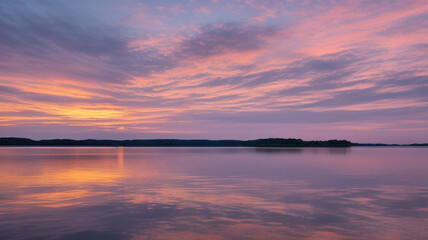 Obraz premium A serene sunset over a calm lake. Wispy clouds are reflected on the water's surface, creating a mirrored effect of the sky.