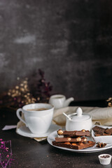 Coffee in a white cup with chocolate, flowers on a dark background, flatlay, chocolate with whole hazelnuts, handmade candies, coffee with milk, exquisite dessert