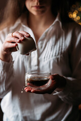 a girl in a white shirt pours milk from a white milk jug into an espresso coffee in a transparent cup, drops of milk in the air, bokeh, coffee with milk, a woman drinks coffee