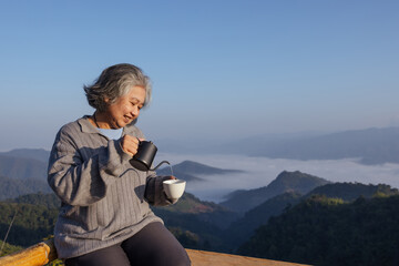 A senior woman enjoys a peaceful morning on a mountain balcony, pouring coffee with a smile. The breathtaking misty valley and blue sky create a serene and relaxing atmosphere in nature