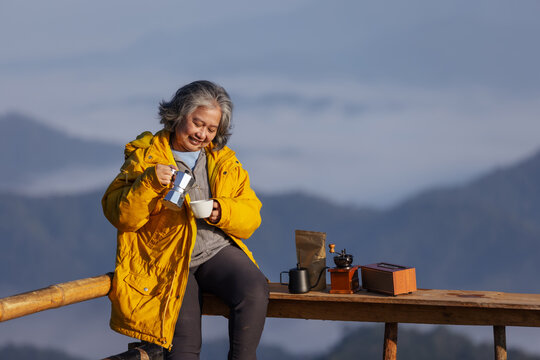 A senior woman in a yellow jacket enjoys a morning coffee on a wooden balcony overlooking misty mountains. With a moka pot and coffee setup beside her, she embraces tranquility, warmth, and nature - Powered by Adobe