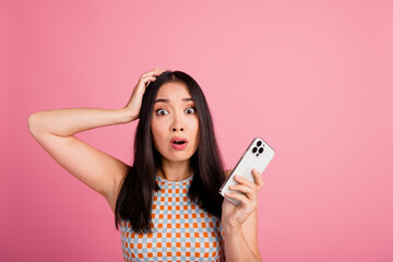Naklejka premium Surprised young Asian woman with smartphone, wearing trendy top, in front of a pink background, experiencing shocking or unexpected news