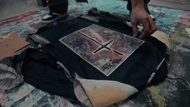 High angle cropped slowmo of hands of unrecognizable artist placing carefully cross shaped pattern on garment working on painting board on floor surrounded by tools and materials