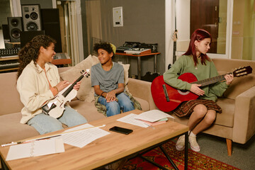 High angle shot of teen boy and girl with bass guitar joking on sofa while their friend strumming...