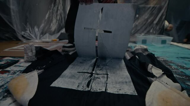 High angle slowmo of young talented Caucasian man removing stencil from black garment leaving white imprint, painting on fabric in workshop with transparent sheeting on walls