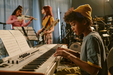 Side view of African American boy concentrated on laying keyboard while his friends chatting in blurred background