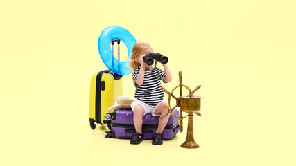 Girl using binoculars near luggage, inflatable ring, and ship wheel, acting as young explorer against yellow studio background. Concept of childhood, vacation, travelling, tourism, marine life