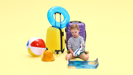 Boy sitting on floor, holding toy airplane over printed ocean image, beach ball and suitcases beside him against yellow studio background. Concept of childhood, vacation, travelling, tourism
