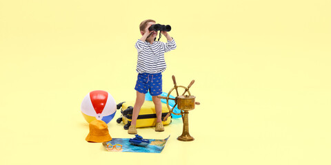Boy looking through binoculars ahead, surrounded by travel props including ship wheel, inflatable ring, printed map against yellow studio background. Childhood, vacation, travelling, tourism concept