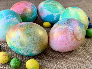 Brightly colored Easter eggs showcase unique patterns in pastel hues of blue, green, yellow, and pink, surrounded by small candy eggs on a natural fabric surface