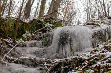 serene winter scene with a small frozen waterfall cascading over rocks covered in snow and moss, surrounded by a bare winter forest. The close-up view highlights the intricate details of the ice forma