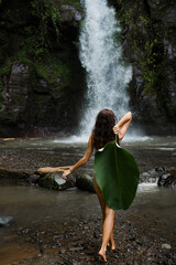 Naked woman covering herself with a banana leaf in front of a tropical waterfall.
