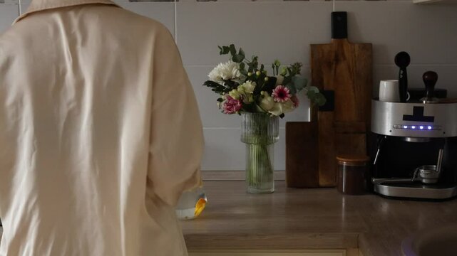 A person arranges flowers in a vase while preparing a drink in a warm kitchen atmosphere.