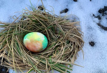 A vibrant Easter egg rests in a nest made of dry grass and surrounded by white snow, indicating the transition from winter to spring