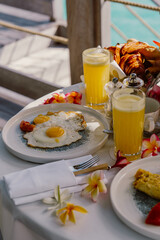 A tropical breakfast setup on a terrace overlooking crystal-clear waters, featuring fresh juice, eggs, pastries, and vibrant red flowers, creating a luxurious and relaxing island dining experience