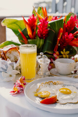 A tropical breakfast setup on a terrace overlooking crystal-clear waters, featuring fresh juice, eggs, pastries, and vibrant red flowers, creating a luxurious and relaxing island dining experience