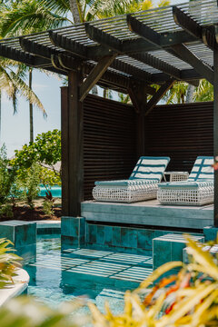 Luxurious poolside cabana with elegant lounge chairs, shaded by a wooden pergola, surrounded by lush tropical greenery. A perfect setting for relaxation and indulgence in a tropical paradise

