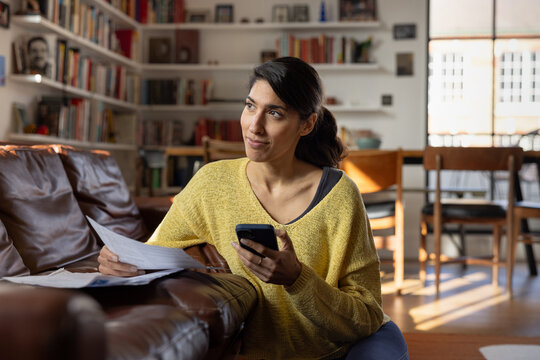 Woman using electronic banking while holding paperwork thoughtfully