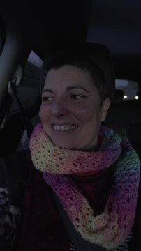 vertical video of woman in car looking out of window, smiling and with light changing on her face. She is wearing a colourful scarf