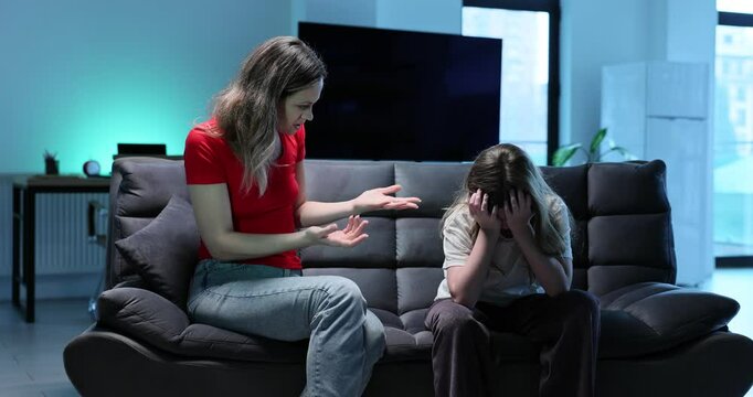 Emotional mother scolds crying daughter sitting on sofa in living room. Woman displeased by girl behavior and school marks conflict slow motion
