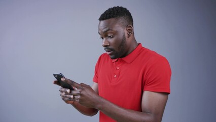 Good-looking young bearded Afro-American man actively uses his smartphone for texting messages on grey background, is surprised, cutely smiles. Communication, being online, chatting. Male portrait