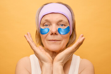 A senior woman enjoys a moment of self-care applying blue eye patches while smiling gently. She is dressed in a simple tank top, showcasing a radiant mood and relaxed environment