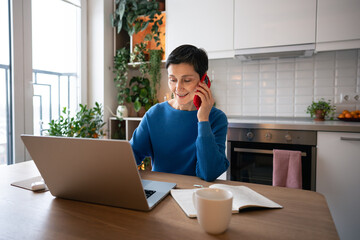 Confident mature woman smiling while talking on smartphone and working on laptop at home. Glad...
