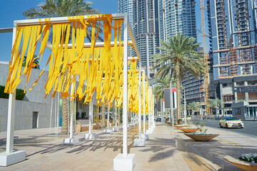 A vibrant urban scene featuring yellow ribbons hanging from a modern structure. Palm trees line the street, with tall buildings under construction in the background.