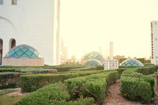 A serene garden with geometric glass domes and lush green hedges, set against a modern architectural building. The sun casts a warm glow over the scene, creating a peaceful atmosphere. - Powered by Adobe
