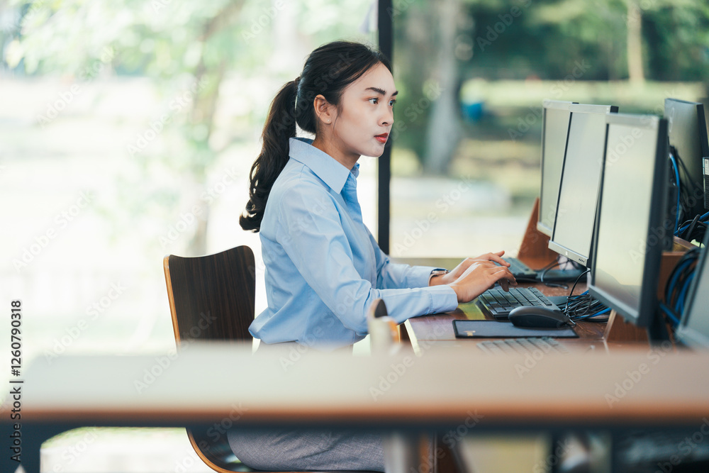 Poster asian businesswoman sitting at her desk working on desktop computer in modern office with multiple m