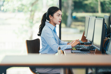 Asian businesswoman sitting at her desk working on desktop computer in modern office with multiple monitors, she is focused on her work and typing on keyboard