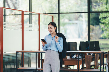 Young Asian businesswoman wearing blue shirt and gray pants, holding jacket over shoulder and using smartphone in modern office with computers in background