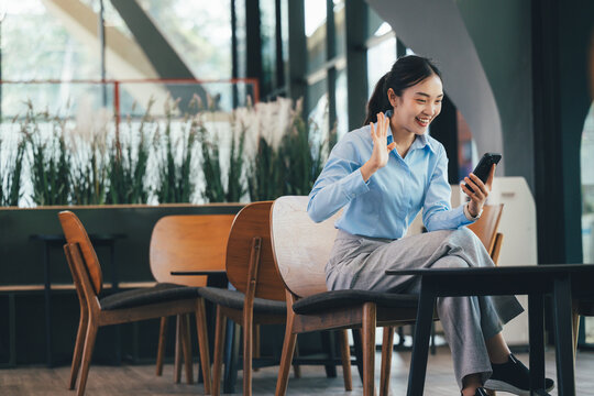 Professional Asian businesswoman waving during video call, sitting in contemporary office cafeteria, connecting remotely with colleagues