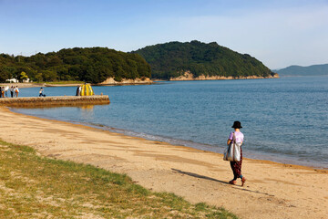 A young lady walking on the sandy beach in Naoshima island & tourists taking photos of Kusama Yayoi's famous artwork, the Yellow Pumpkin, on a jetty during Setouchi Triennale Festival in Kagawa, Japan