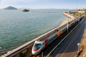 A Shiokaze Express Train travels on JR Yosan Line along the beautiful coast with Tsushima Jinja Shrine on an offshore island connected by a bridge to the beach, in Minocho Omi, Kagawa, Shikoku, Japan