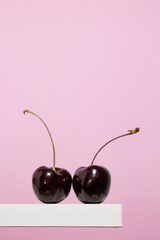 Vertical image of a pair of cherries on a white wooden shelf on a pink background, small red round fruit