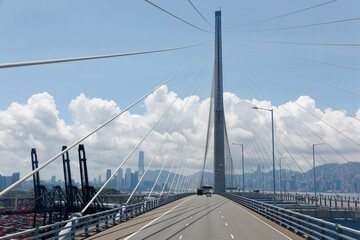 View from a car driving under the tower of Stonecutters Bridge, a cable-stayed bridge spanning Rambler Channel in Hong Kong with cranes on the container terminal & skyscrapers standing under sunny sky