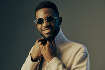 Fototapeta premium Stylish young African man in sunglasses smiles confidently, wearing a fashionable coat against a neutral background, showcasing contemporary fashion and joy