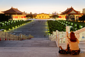 Fototapeta premium A tourist sitting on the stairway to Chiang Kai Shek Memorial Hall overlooking Liberty Square flanked by National Theater & Concert Hall and the majestic archway illuminated before nightfall in Taipei