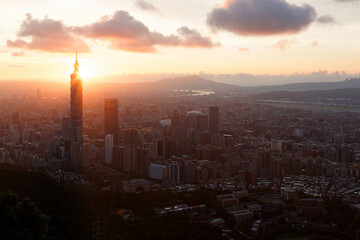 Aerial view of Downtown Taipei at sunset, with prominent 101 Tower standing among skyscrapers in Xinyi District and Tamsui River meandering by distant mountains under golden twilight sky in background