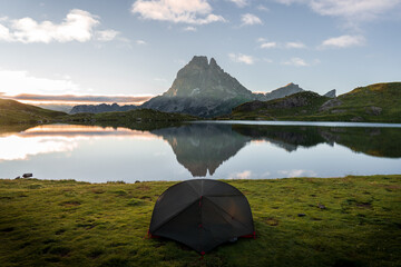 Green tent in front of a lake in which the mountains are reflected at dawn