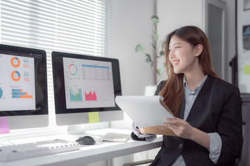 Young professional woman reviewing documents and analyzing data displayed on computer monitors, engaged in business planning and financial analysis in a modern office setting