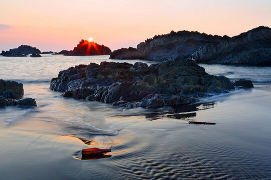 A photographer taking photos of sunrise at a rocky beach with golden sunlight reflected on the water, sea waves rushing upon the shore & Guishan Island on horizon at Wai'ao Coast, Ilan (Yilan), Taiwan - Powered by Adobe